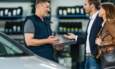 Mecânico de autocenter conversando com um casal de clientes ao lado de um carro, com prateleiras de pneus ao fundo